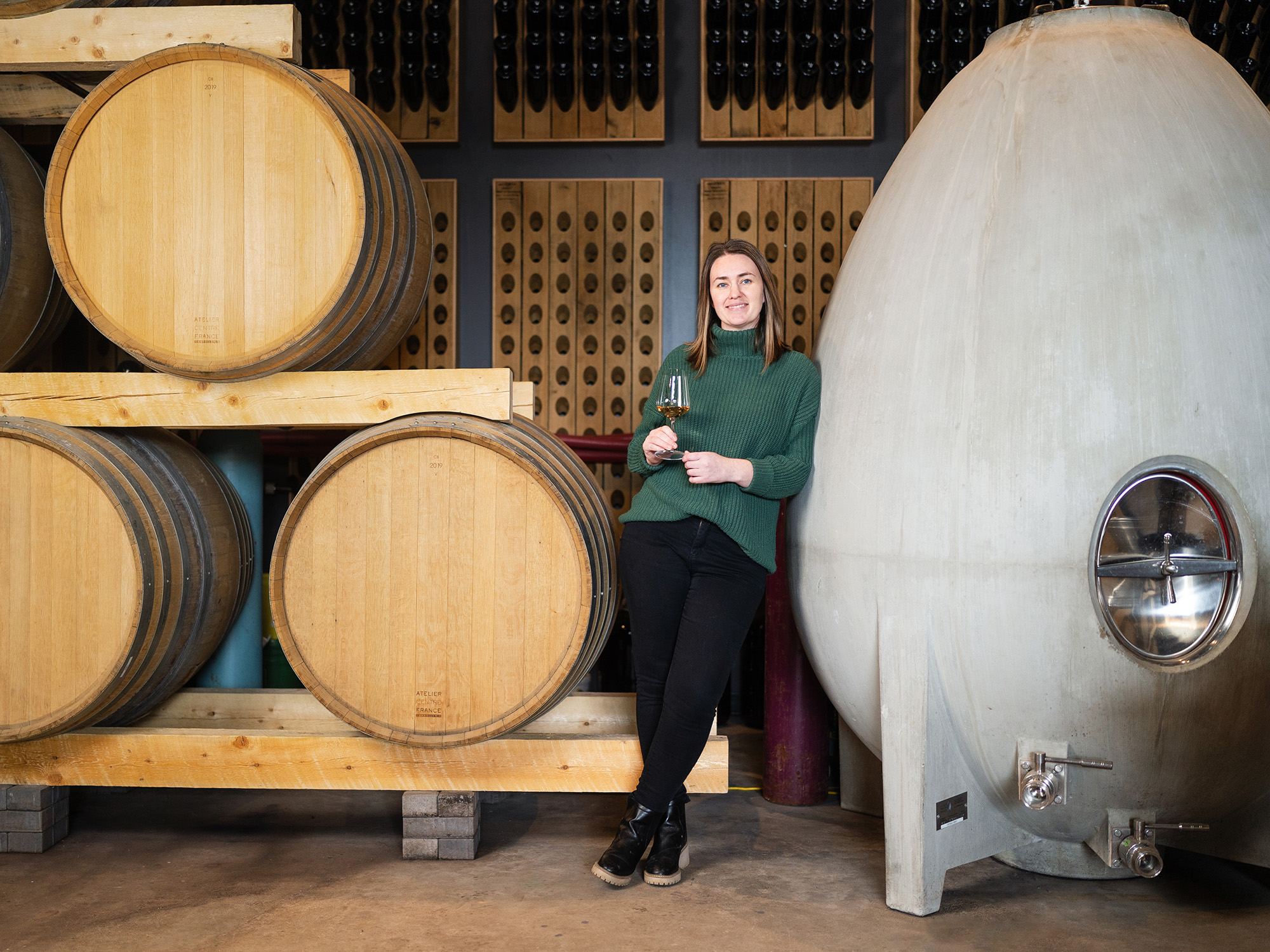 A woman in a green sweater and black pants standing with a glass of wine beside wooden wine barrels.