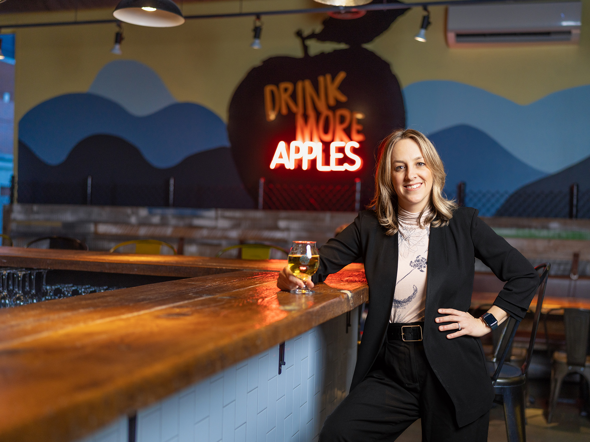 A woman in a black business suit holds a a glass of cider and is standing beside the bar at Chain Yard Urban Cidery & Taproom